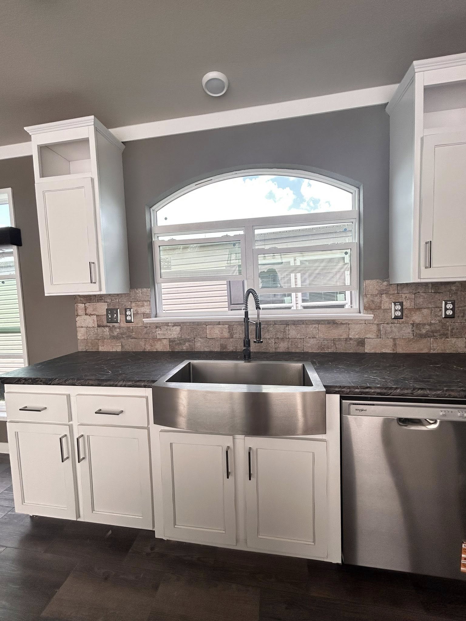 Modern kitchen with white cabinets, stainless steel farmhouse sink, and dishwasher. Large arched window above sink and stone tile backsplash.