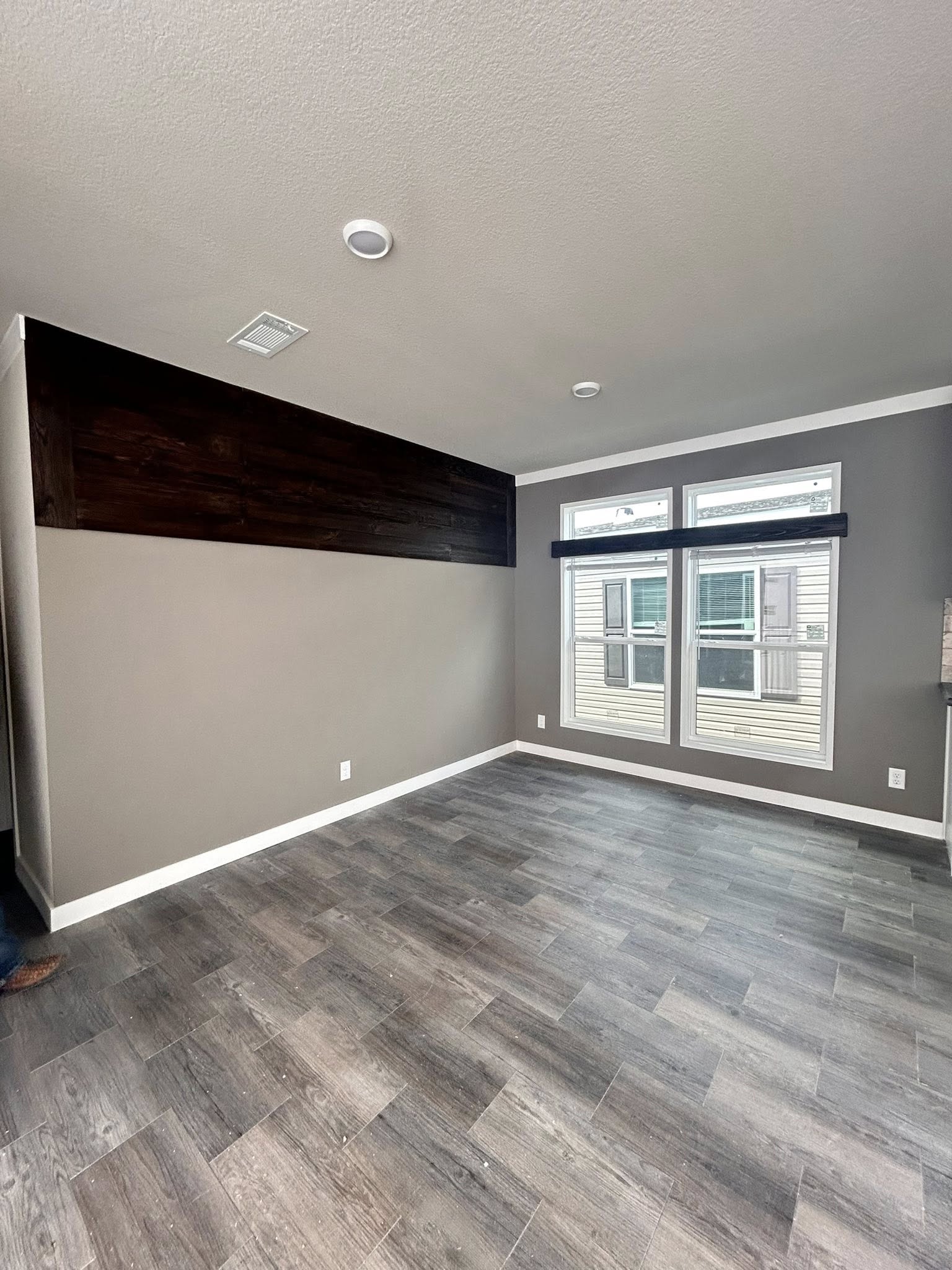 Empty room with gray walls and dark wood paneling. Two large windows with black blinds let in natural light. Gray wood-style flooring adds a modern touch.