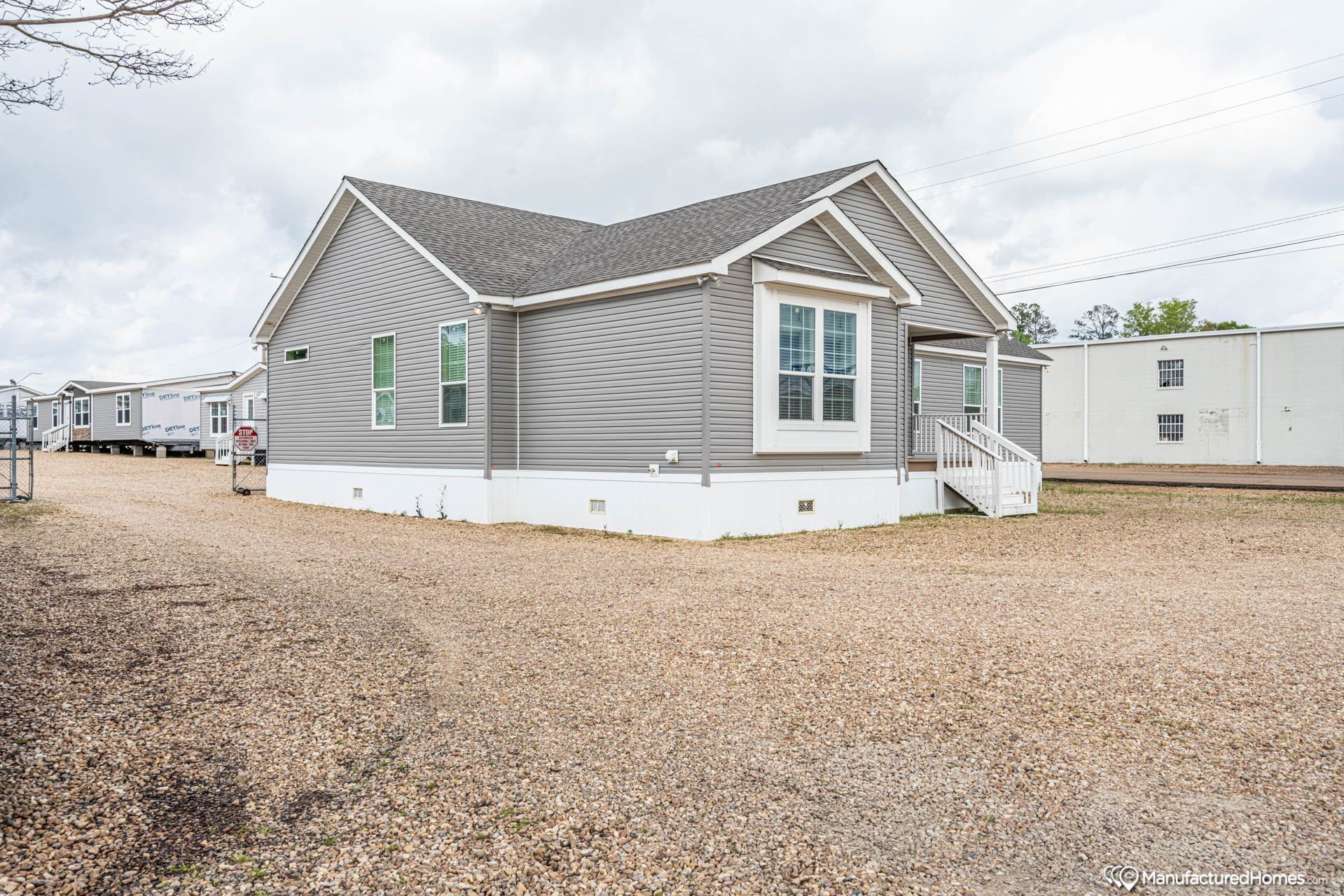 A grey manufactured home with white trim sits on a gravel lot under a cloudy sky. The home's simple design and windows convey a practical tone.