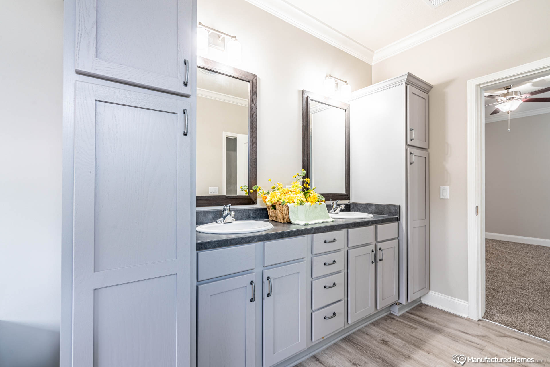 Modern bathroom with gray double vanity and black countertops, featuring two mirrors and a basket of yellow flowers. Soft lighting, serene and tidy.