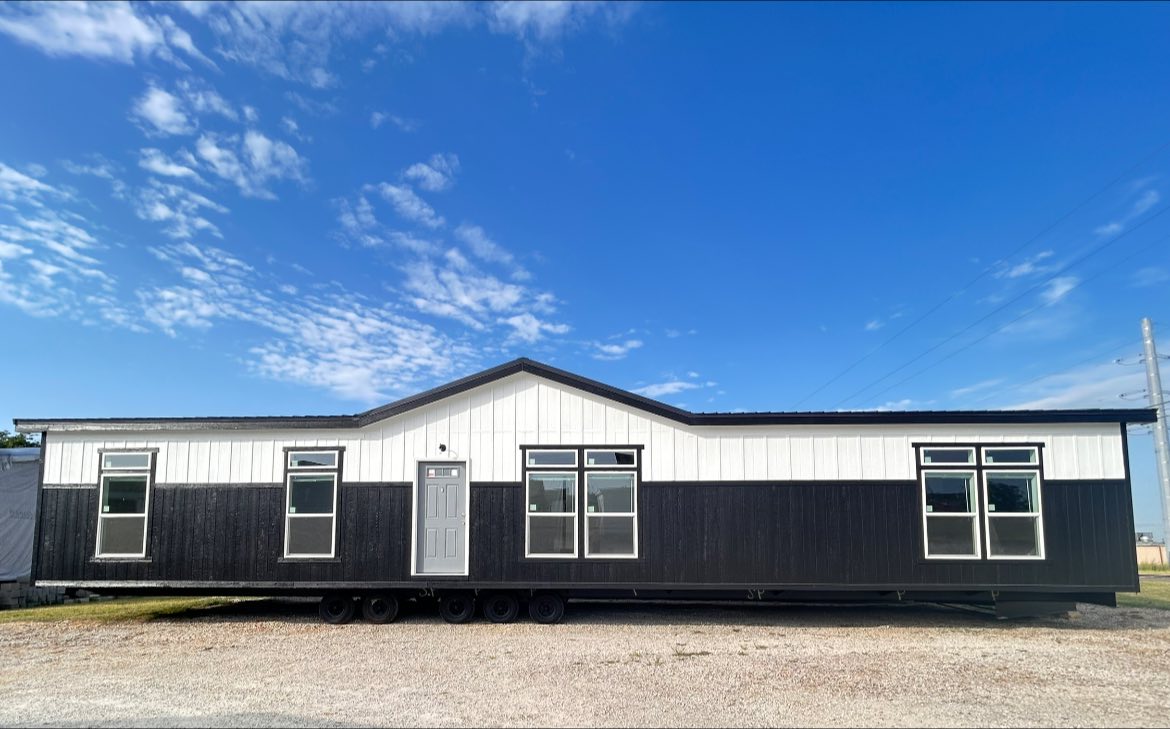 Modern black and white mobile home under a clear blue sky. The house features large windows and a central gray door, conveying a sleek, contemporary look.