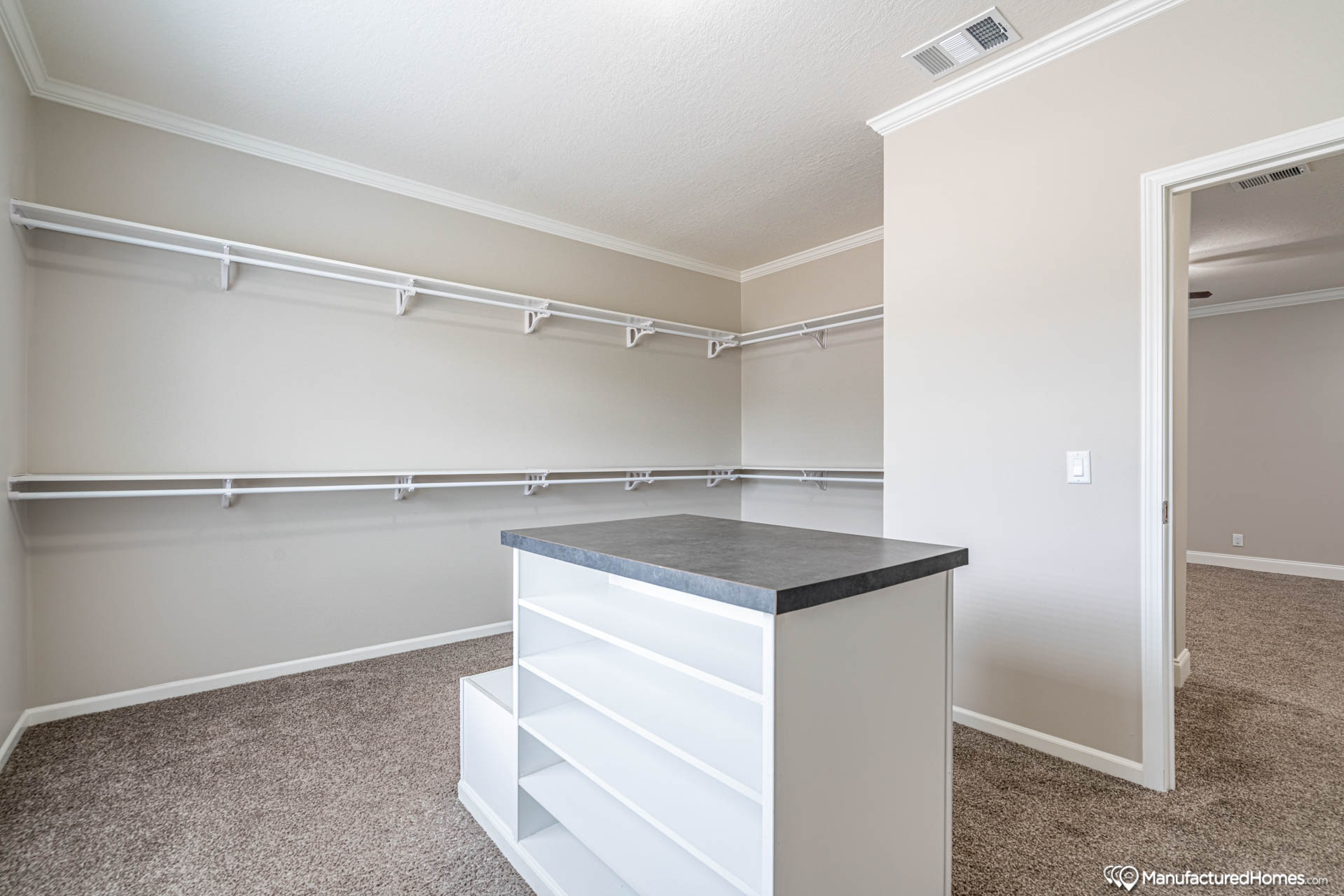 Spacious walk-in closet with beige walls and carpet, featuring an island with open shelves. White shelving lines the walls, creating an organized and clean look.