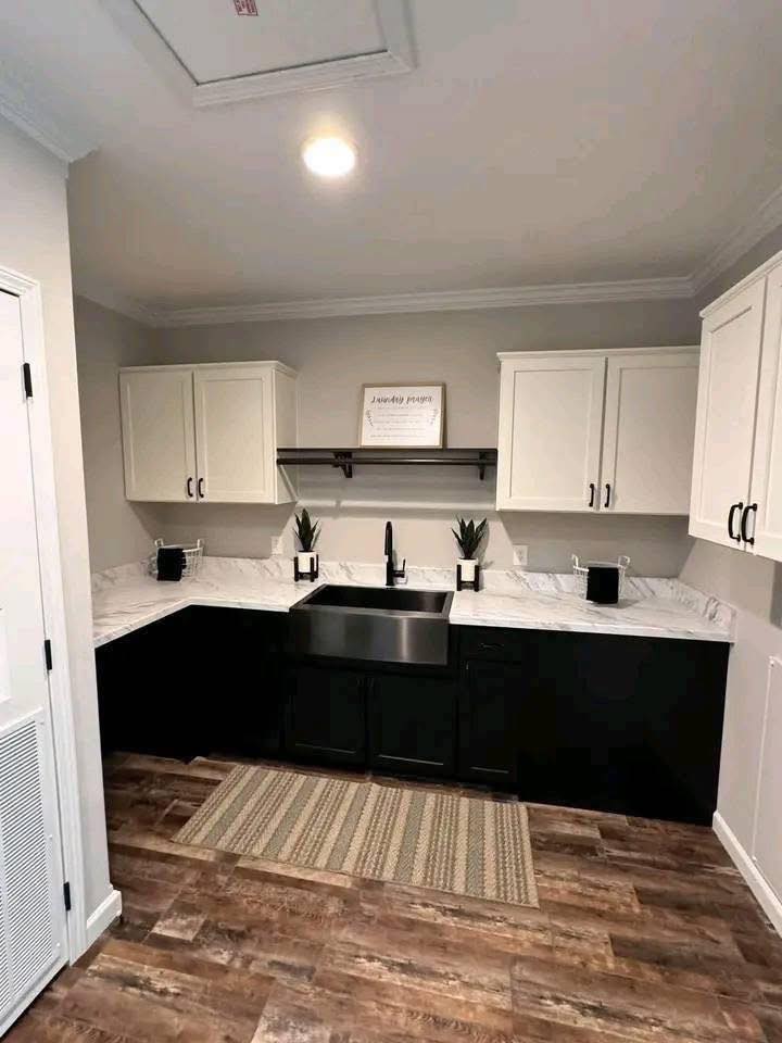 A modern kitchen with white upper cabinets, black lower cabinets, and a stainless steel sink. Marble countertops, potted plants, and wooden flooring add warmth.