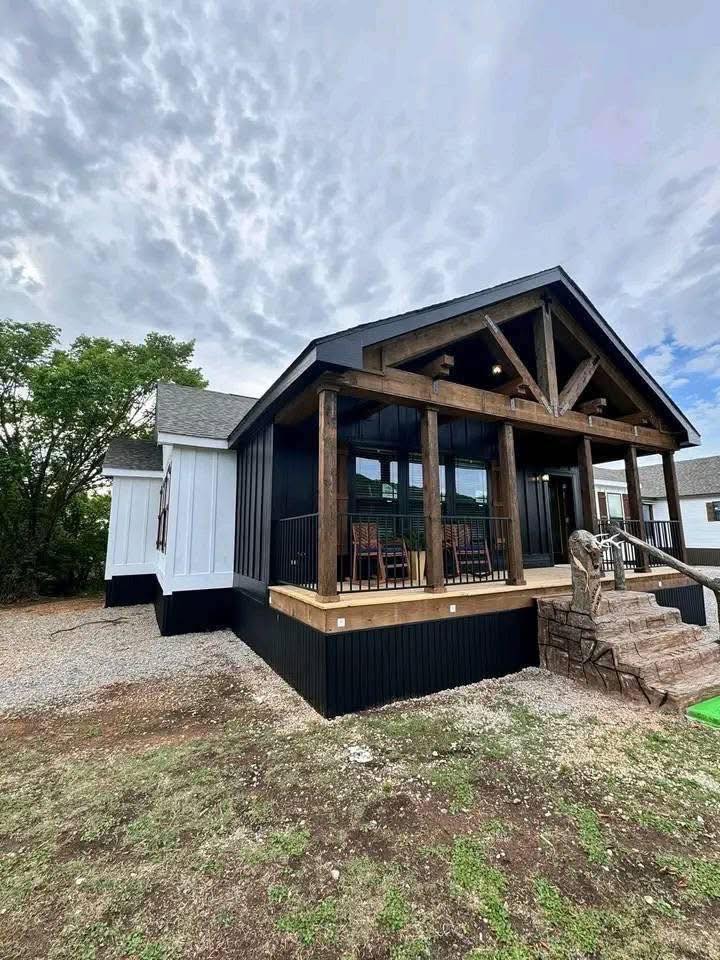 Rustic modern house with wooden beams and a large porch featuring rocking chairs. Cloudy sky, lush greenery in the background, creating a cozy, inviting atmosphere.