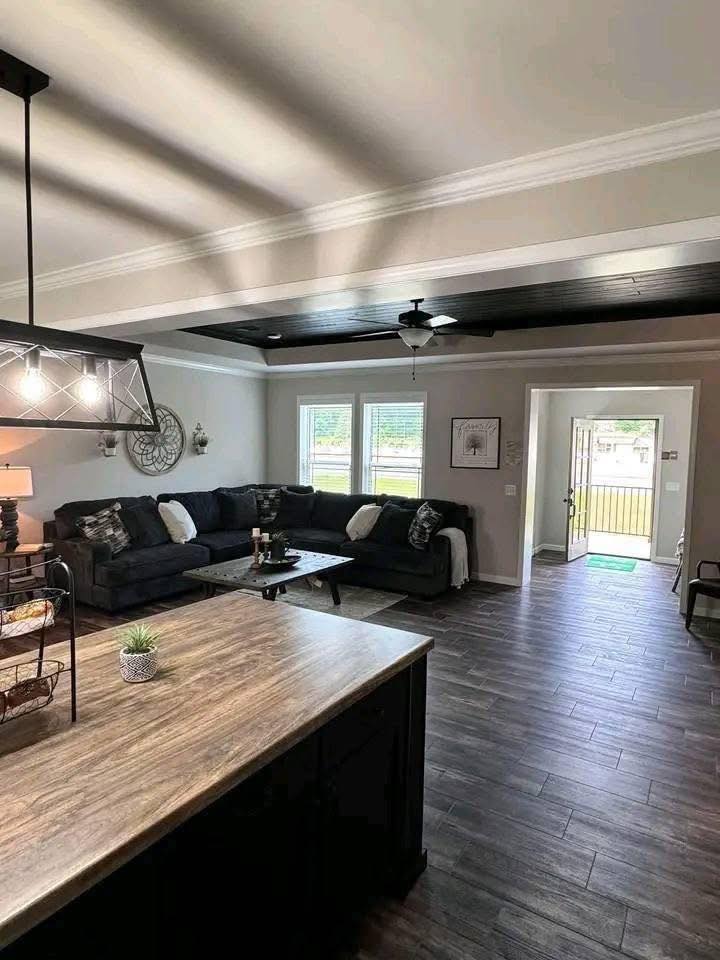 Modern living room with dark wood floors, a large black sectional sofa, and a wooden coffee table. Sunlight streams through two windows, creating a cozy atmosphere.