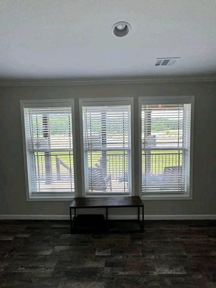 A room with three large windows featuring white blinds letting in natural light, a wooden bench beneath them, set against a calm, neutral wall.