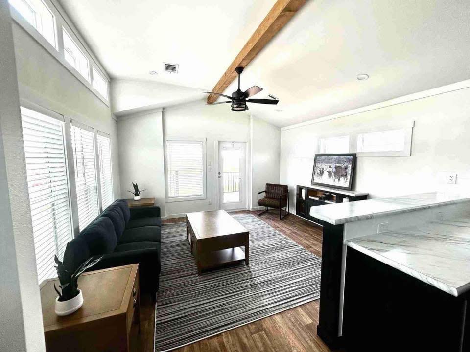 A bright, modern living room with a high ceiling, exposed wooden beam, and large windows. Features a black sofa, striped rug, wooden coffee table, and ceiling fan.
