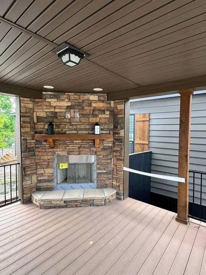 Covered patio with brown wooden ceiling and floor, featuring a stone fireplace. A wooden mantel holds two decorative jars, creating a cozy atmosphere.