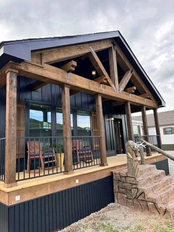 Rustic modern cabin with a wooden porch, vaulted roof, and stone steps. Two wooden chairs sit on the porch, set against a cloudy sky backdrop.