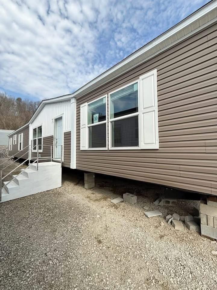 A brown manufactured home elevated on cinder blocks with metal stairs leads to a white door, set against a cloudy blue sky, evoking a rural setting.