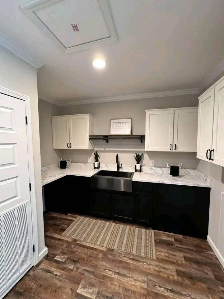 A modern kitchen with white upper cabinets and black lower cabinets. Features a black sink, white countertop, two plants, wood floor, and a light gray wall. Cozy and stylish.