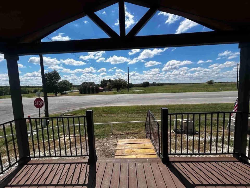View from a porch with wooden steps and black railings, overlooking a quiet rural road. A stop sign is visible under a clear sky with fluffy clouds.