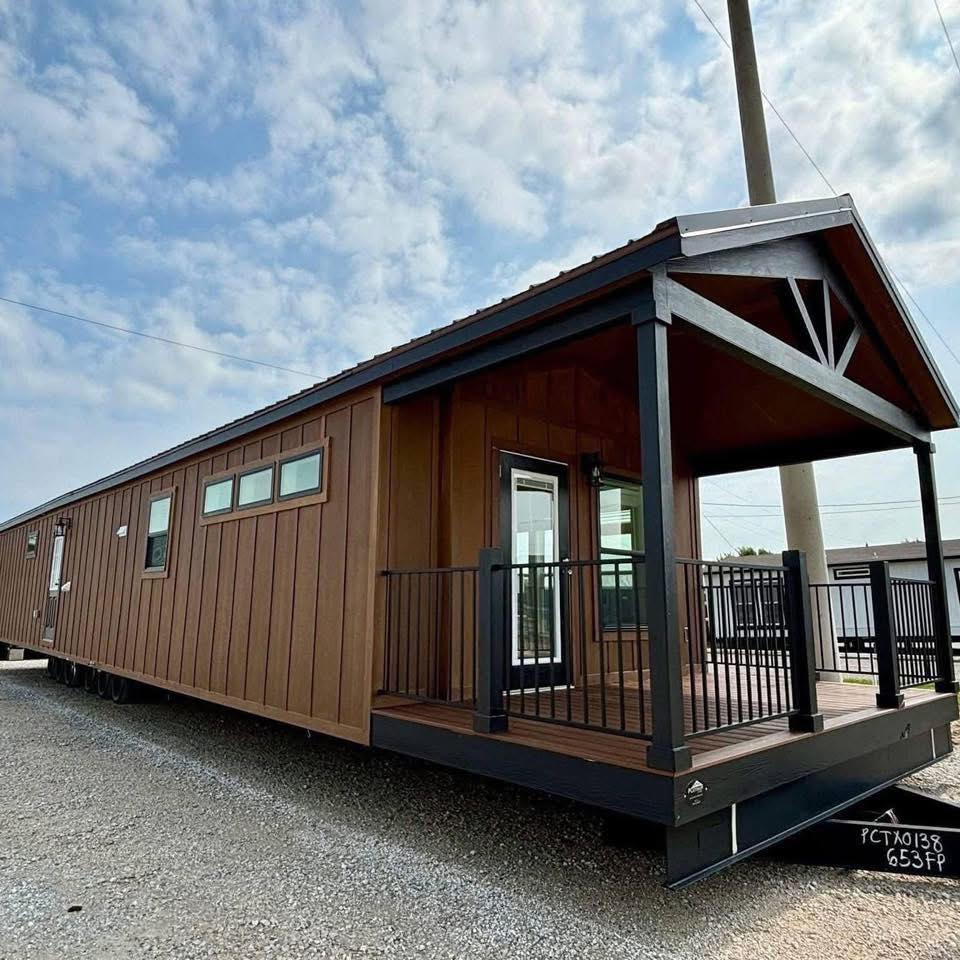 A brown tiny home with a gabled roof sits on gravel, featuring a porch with black railings. The sky is partly cloudy, adding a serene backdrop.A brown tiny home with a gabled roof sits on gravel, featuring a porch with black railings. The sky is partly cloudy, adding a serene backdrop.