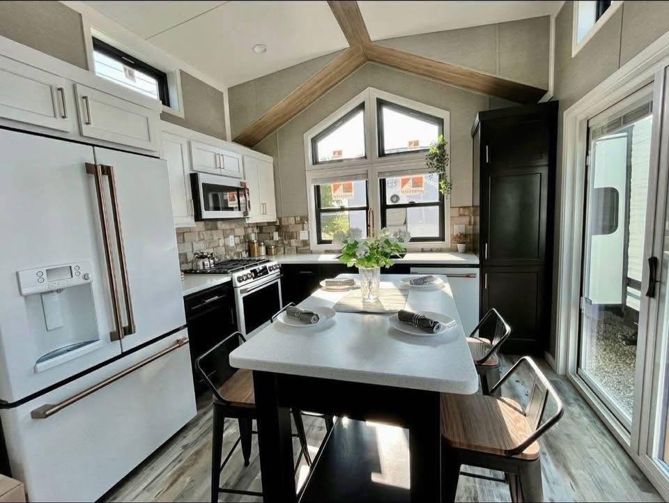 Modern kitchen with high ceiling and wooden beams, featuring a central island with stools, white cabinets, large windows, and bright natural light.