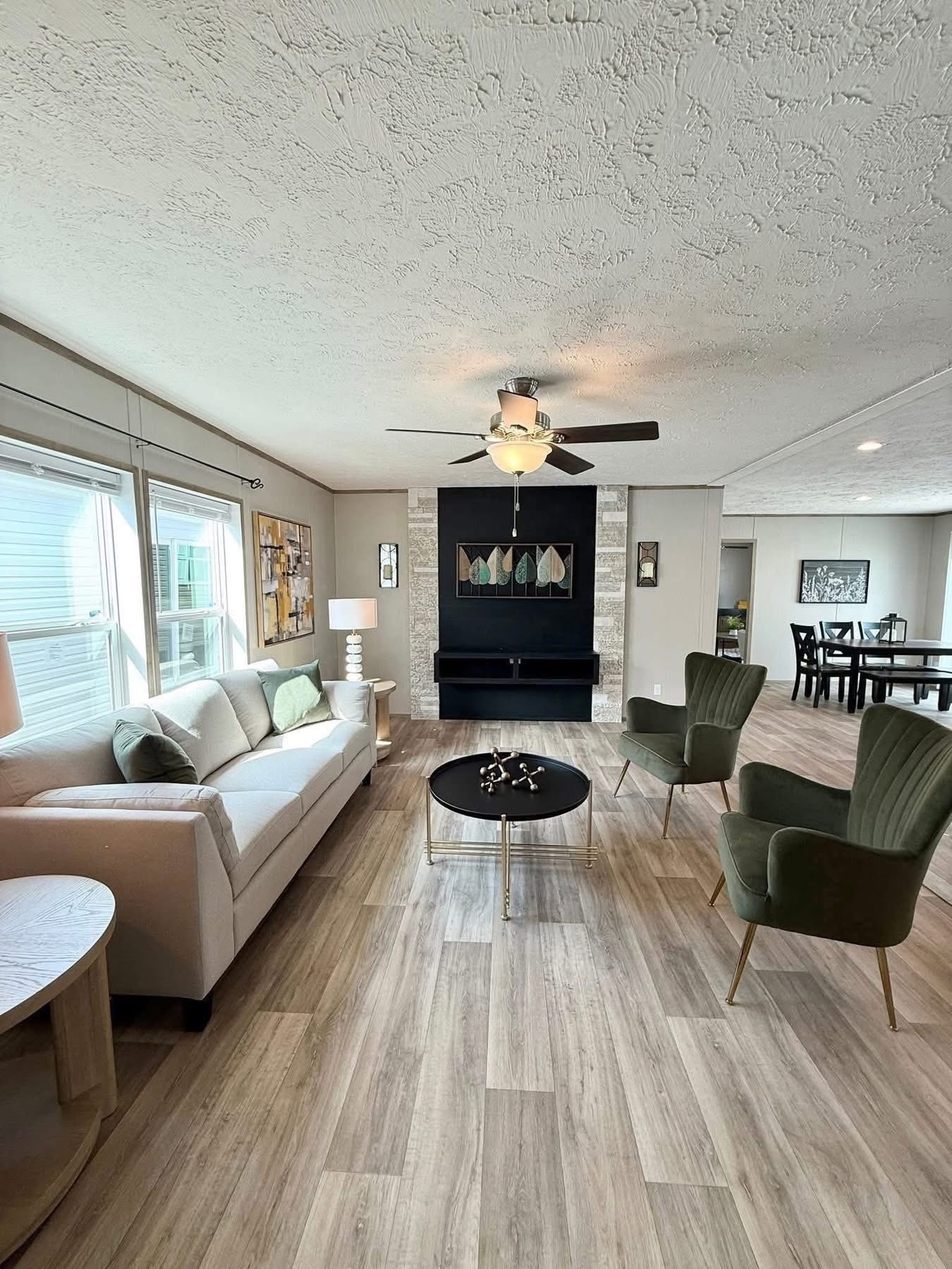 Modern living room with light wood flooring, a beige sofa, two green chairs, and a black round table. A ceiling fan and artwork create a cozy ambiance.