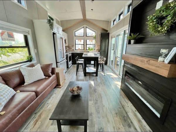 Modern tiny home interior with a cozy living space. Features a brown leather sofa, wood accents, and a sleek kitchen with ample natural light.