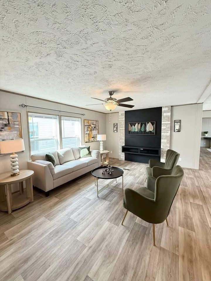 Spacious living room with a light beige sofa, green armchair, and a round coffee table. Large windows, ceiling fan, and modern wall art create a cozy atmosphere.