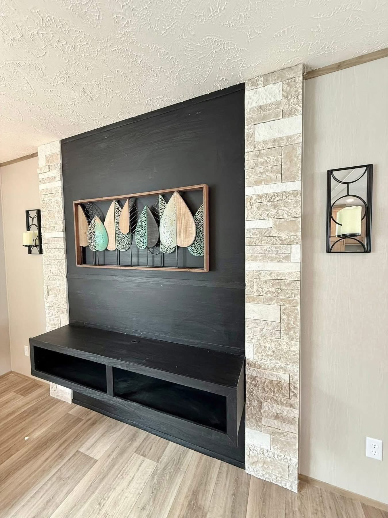 Modern living room with a black accent wall featuring colorful leaf art. Flanked by stone-textured borders and a sleek black shelf below. Elegant and serene.