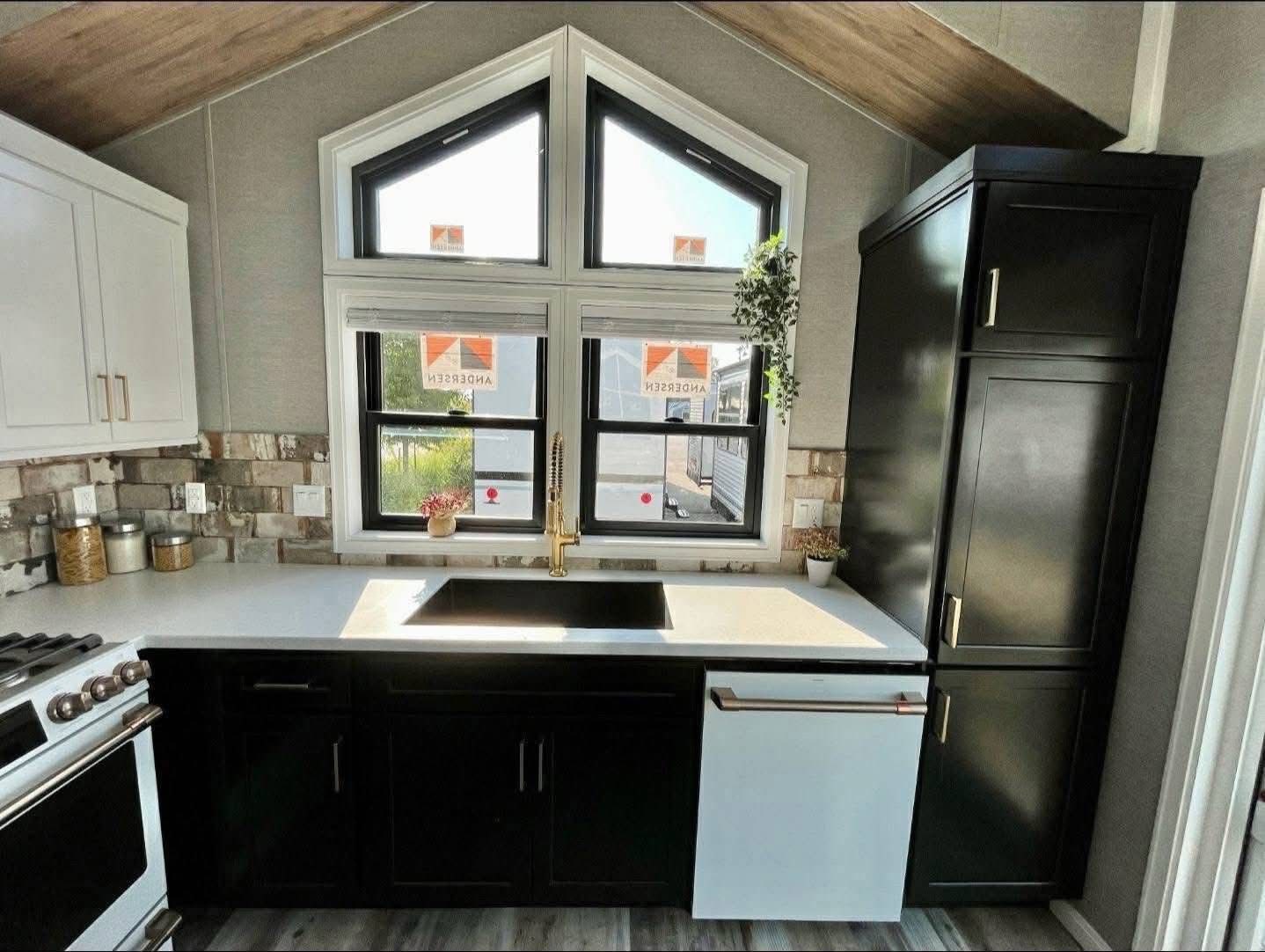 Modern kitchen with a vaulted ceiling, featuring a triangular window over a white countertop and black cabinets. Greenery and brick accents add warmth.