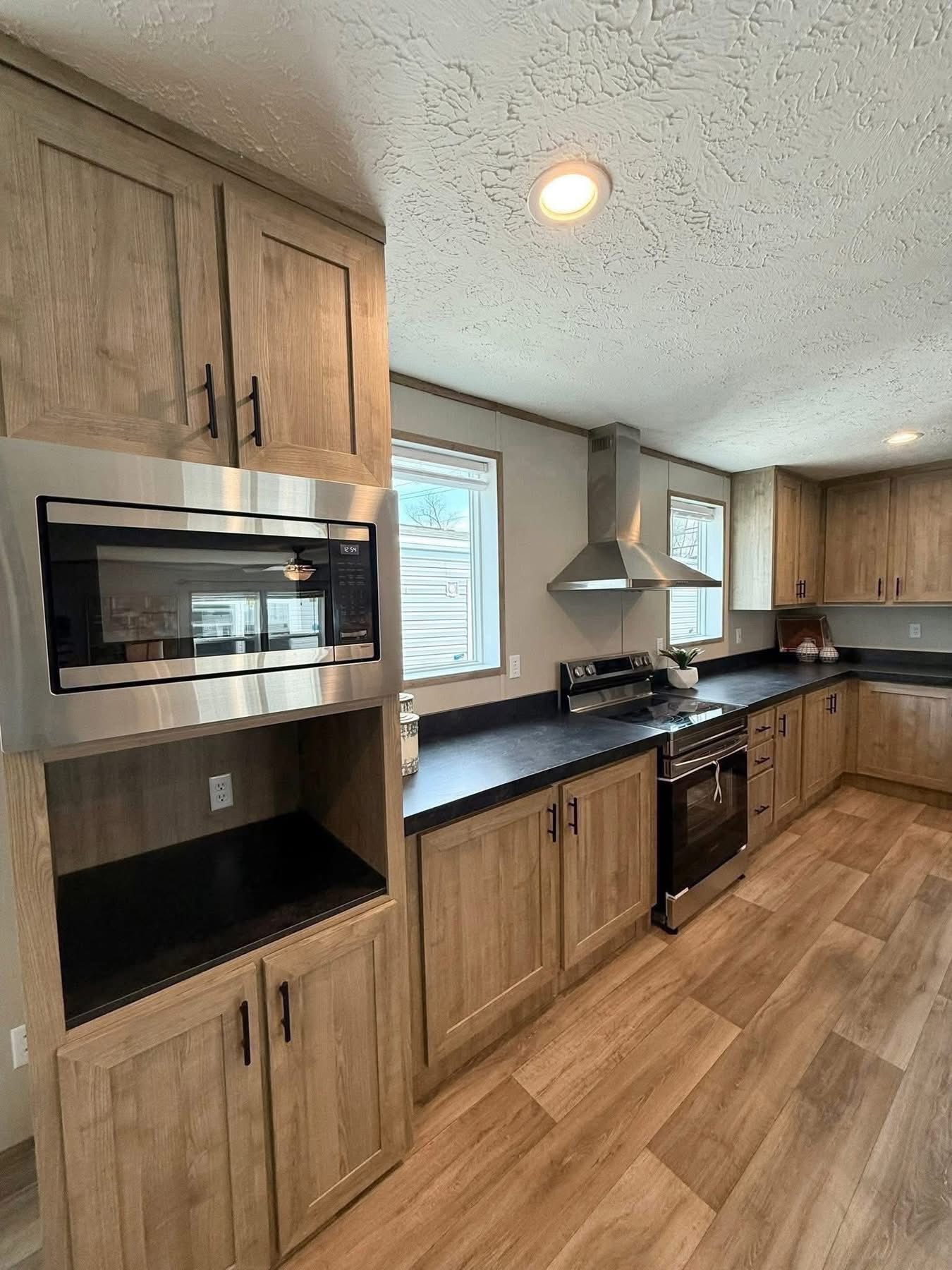 Modern kitchen with wood cabinets, black countertops, stainless steel appliances, and light wood flooring. Bright and spacious with two windows.