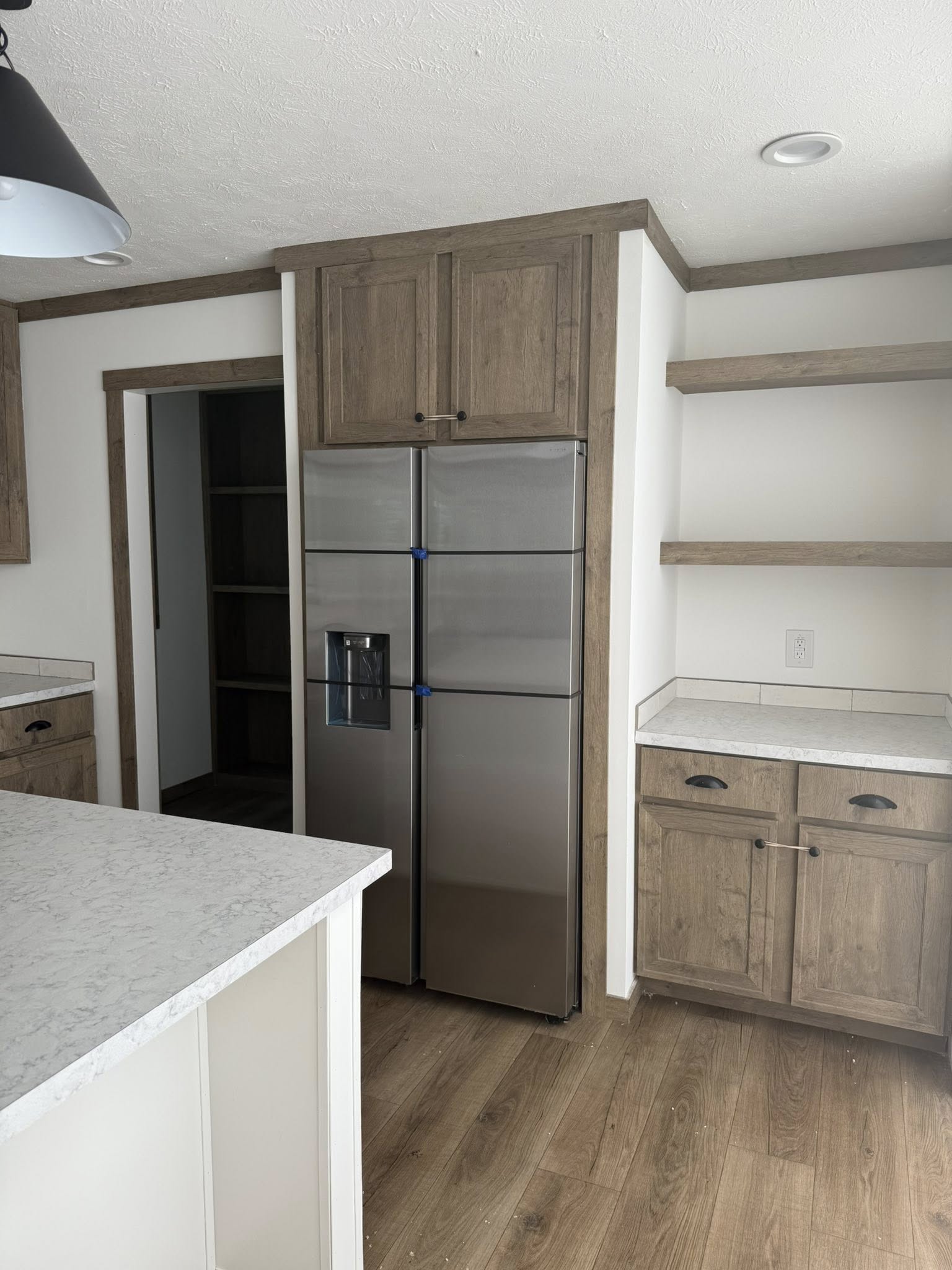 Modern kitchen with stainless steel fridge, light wood cabinets, and marble countertops. Open shelves and a pantry door add a spacious feel.
