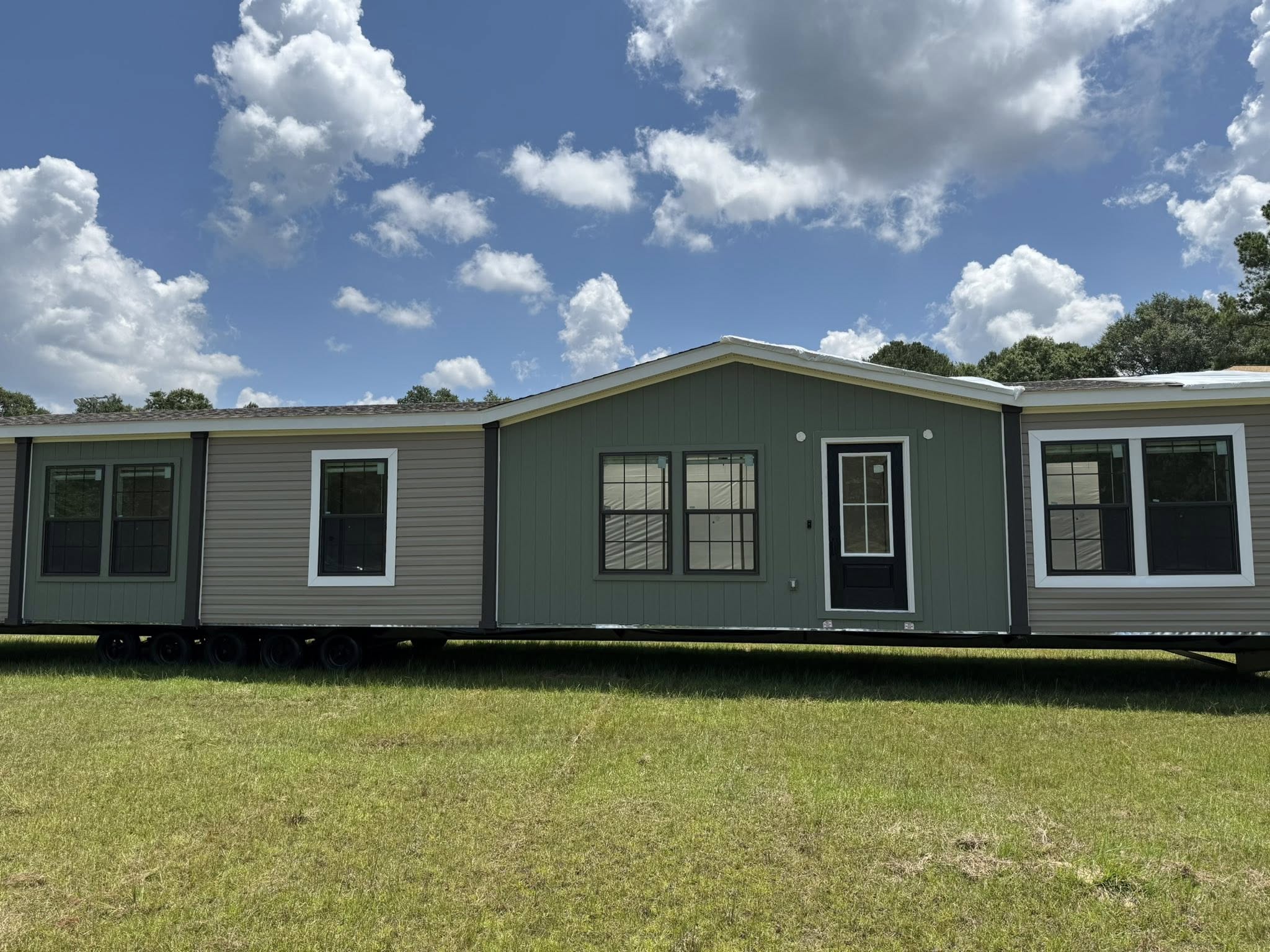 A single-story manufactured home with two-tone siding sits on a grassy field under a partly cloudy sky. It features multiple large windows and a central door.