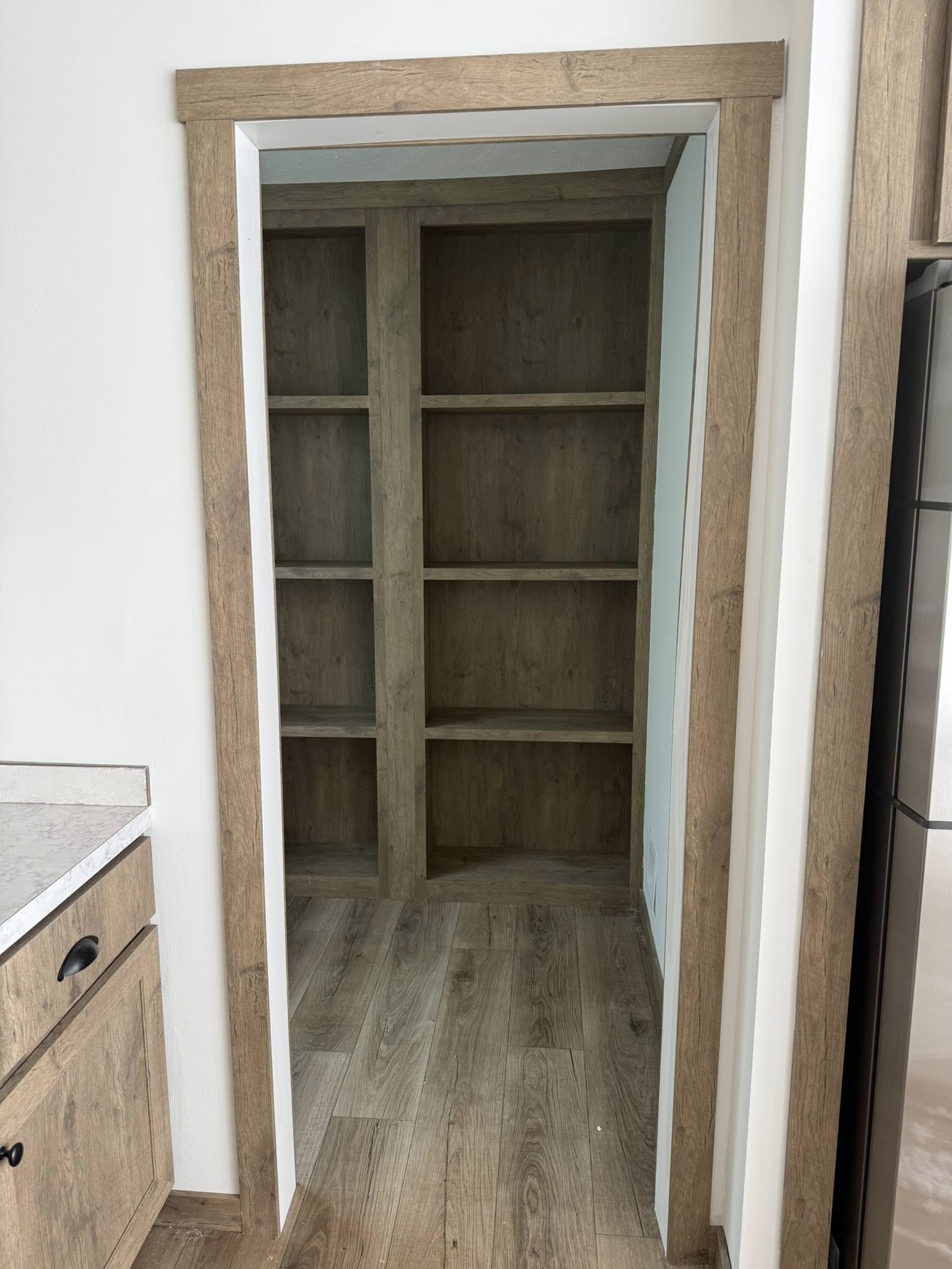 Wood-themed pantry with empty shelves and hardwood floor, framed by a light-tan doorway. Adjacent is a partial view of a kitchen cabinet.