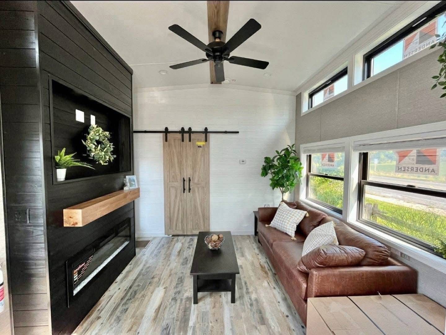 Stylish living room with brown leather sofa, large windows, and a wooden coffee table. Black panel wall with plants, wooden accents, and cozy decor.