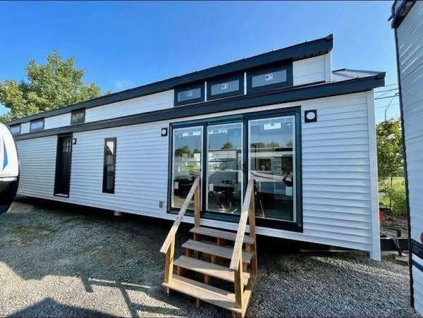 Modern tiny house with white siding and large glass doors, set on gravel. Wooden steps lead to the entrance. Clear blue sky and trees in the background.