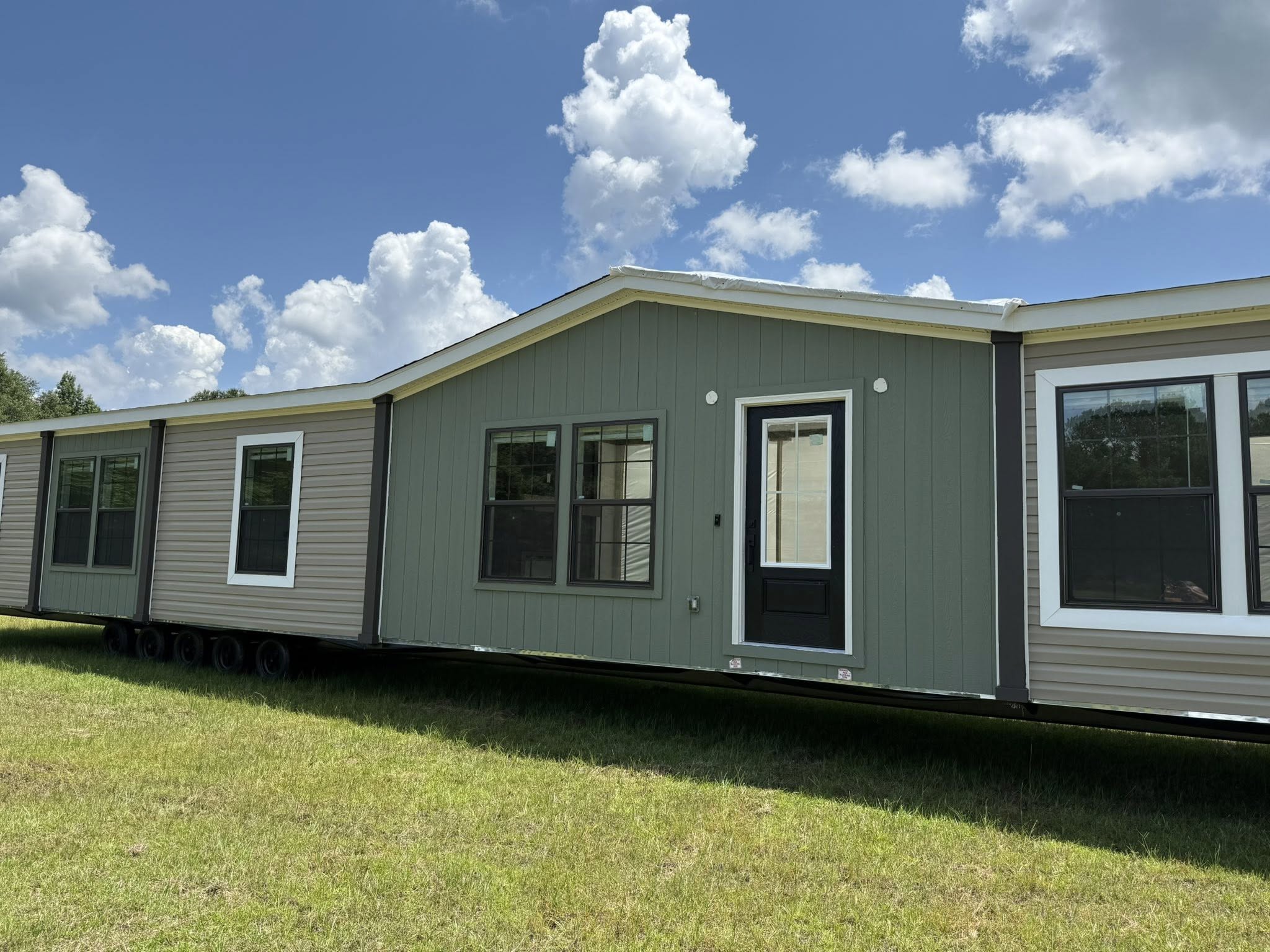 A large manufactured home sits on a grassy field under a blue sky with fluffy clouds. The home has a green and beige exterior, multiple windows, and a central door.