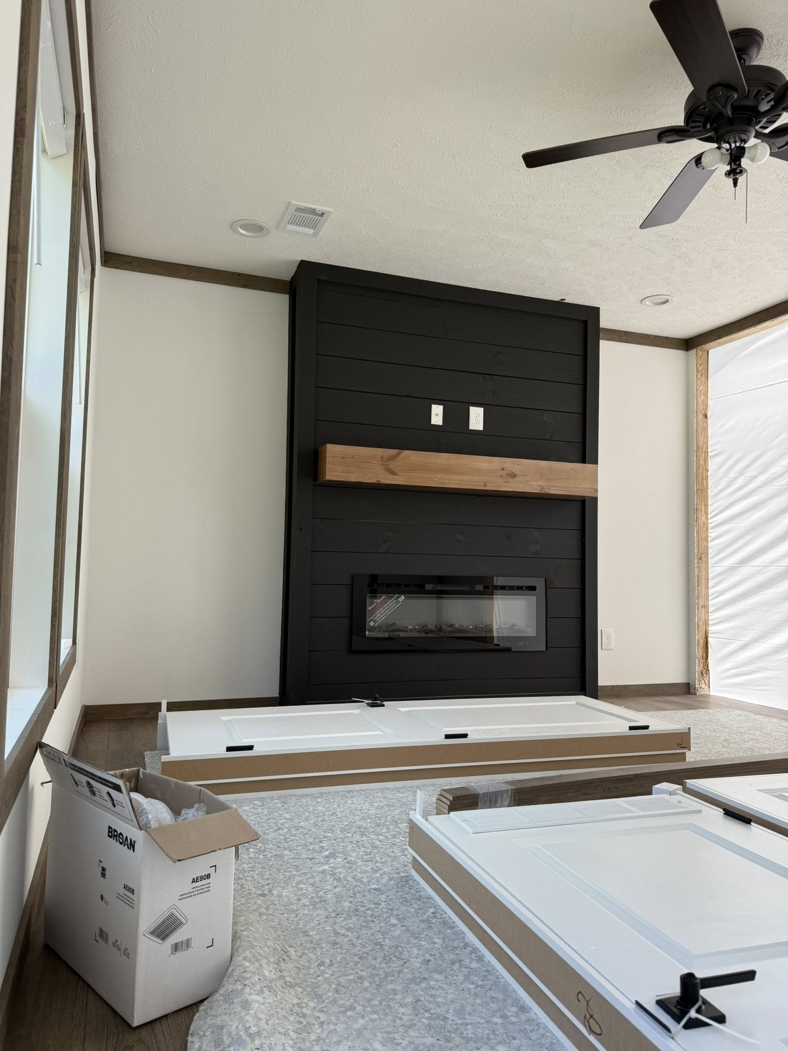 A room under renovation with a black shiplap fireplace wall featuring a wooden mantel. Ceiling fan above and unpacked cabinet doors on the floor convey a work-in-progress feel.