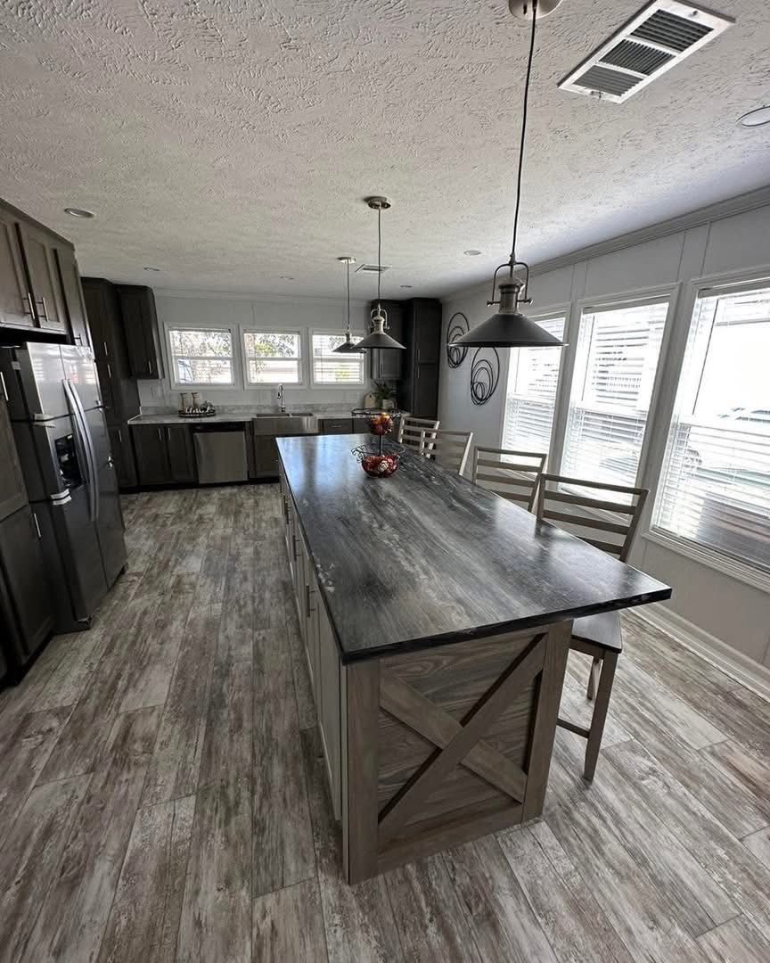 Spacious kitchen with wood flooring, large island with dark countertop, pendant lights, stainless steel appliances, and natural light from multiple windows.