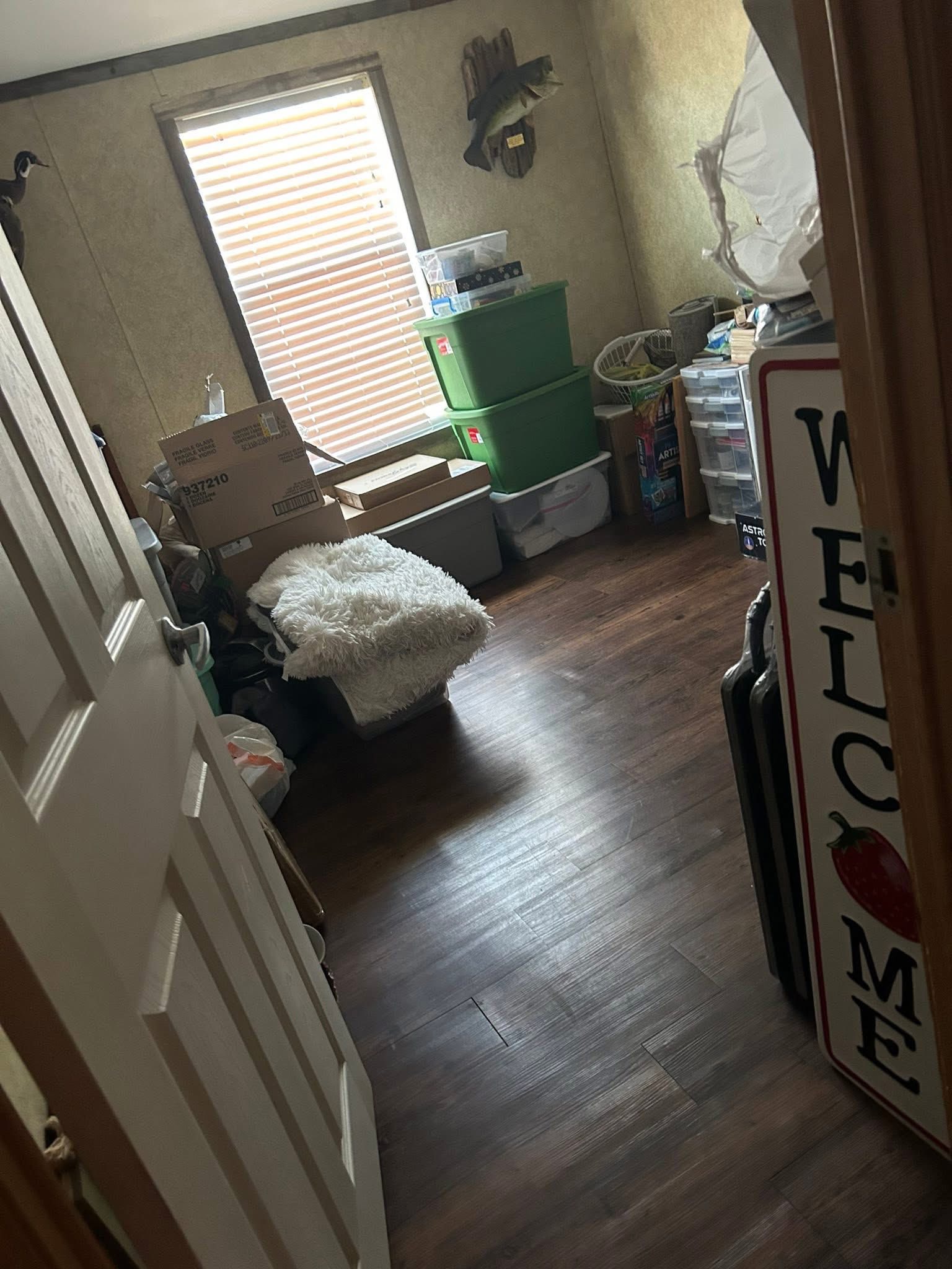 A small cluttered room with boxes, green storage bins, and various household items. A window with blinds lets in dim light, highlighting wooden floors.