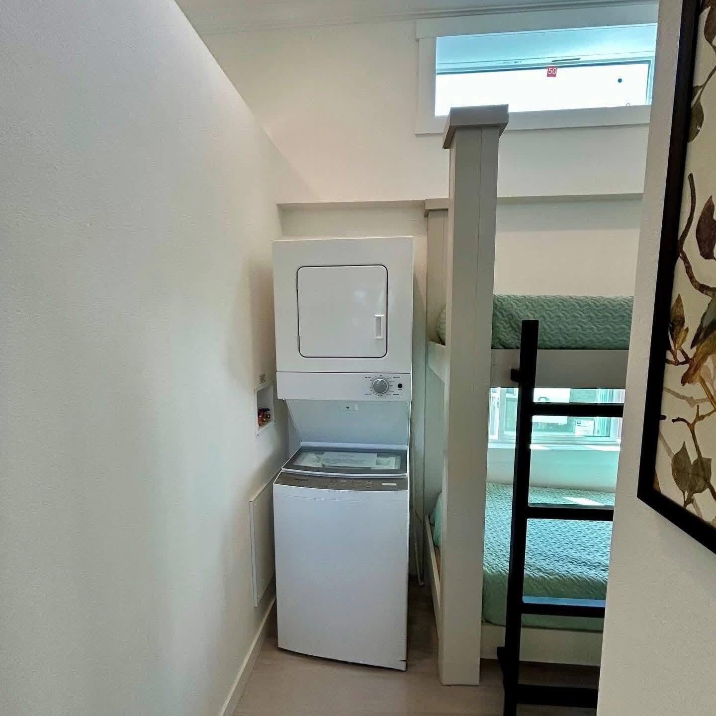 A compact room featuring a stacked washer-dryer next to a bunk bed with teal bedding. Natural light filters through a high window, creating a cozy atmosphere.