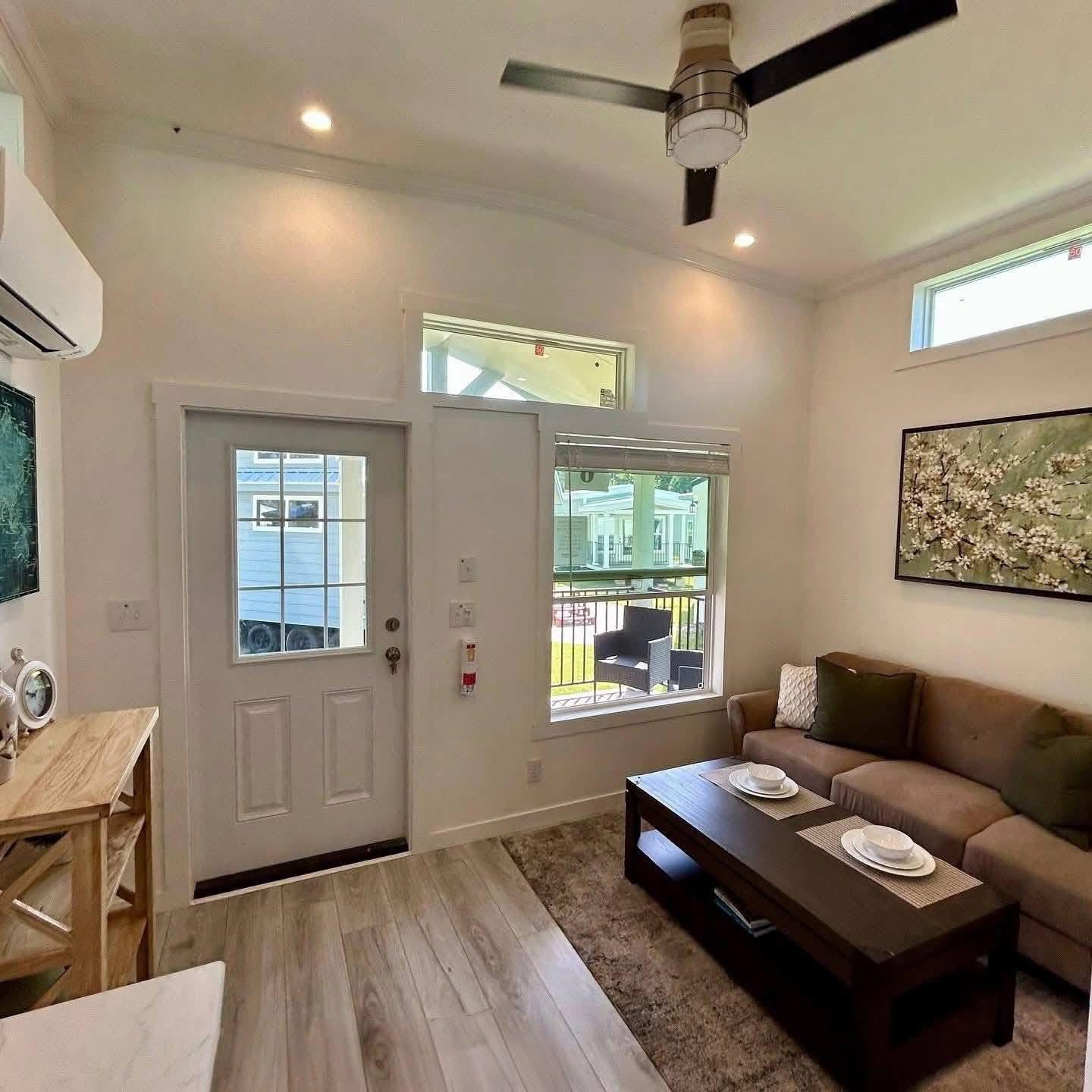 Bright living room with a ceiling fan, light wood flooring, a cozy brown couch, and a wooden coffee table set with white plates. Through the window and glass door, a porch and neighboring houses are visible. A large wall art piece adds a serene touch.