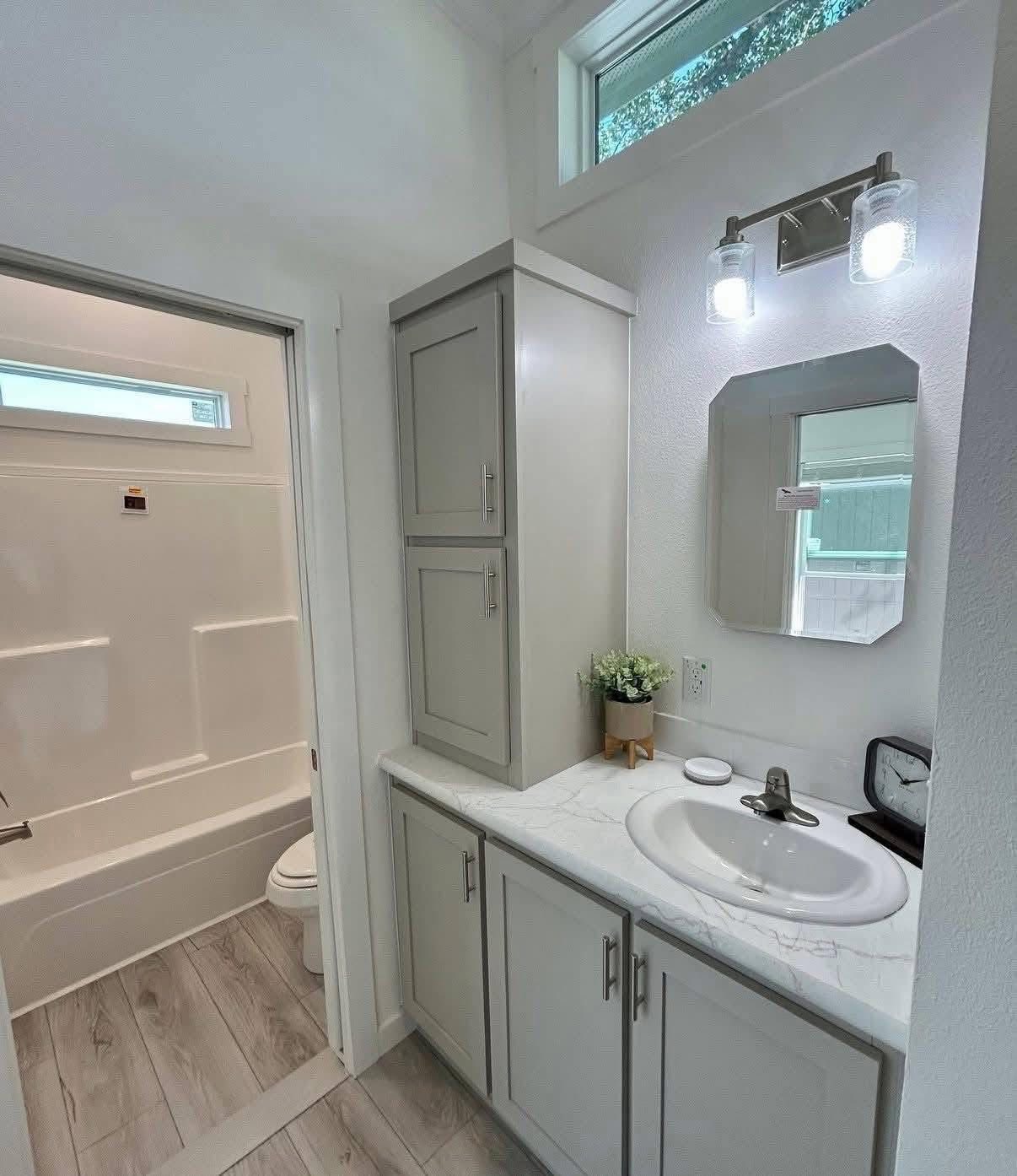 Small, modern bathroom with light gray cabinets, white countertop, and a round sink. Bathtub on the left, mirror, plant, and bright lighting above.