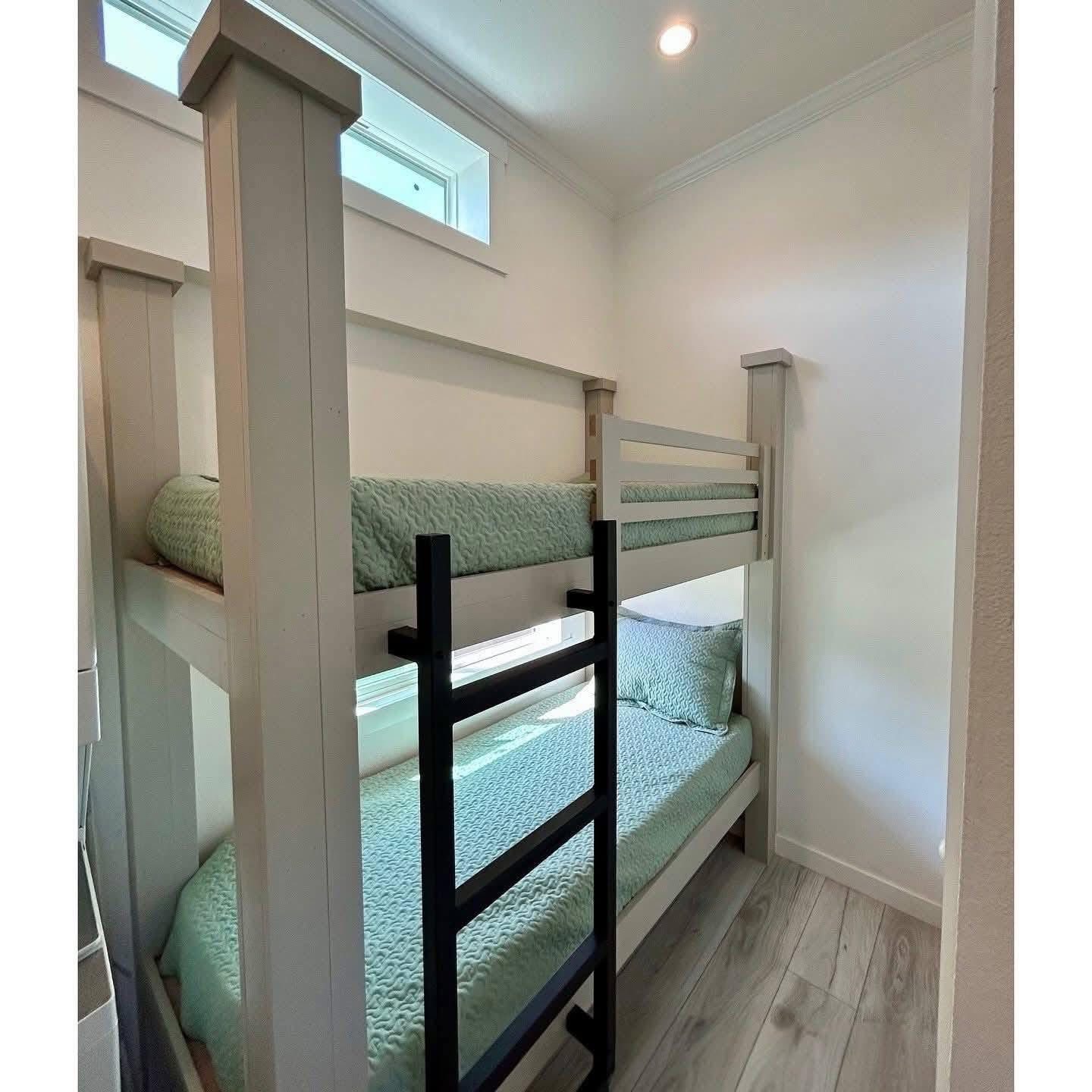 Compact room with white walls featuring a wooden bunk bed, mint-colored bedding, and a black ladder. A narrow window provides soft light. Cozy and minimalist.