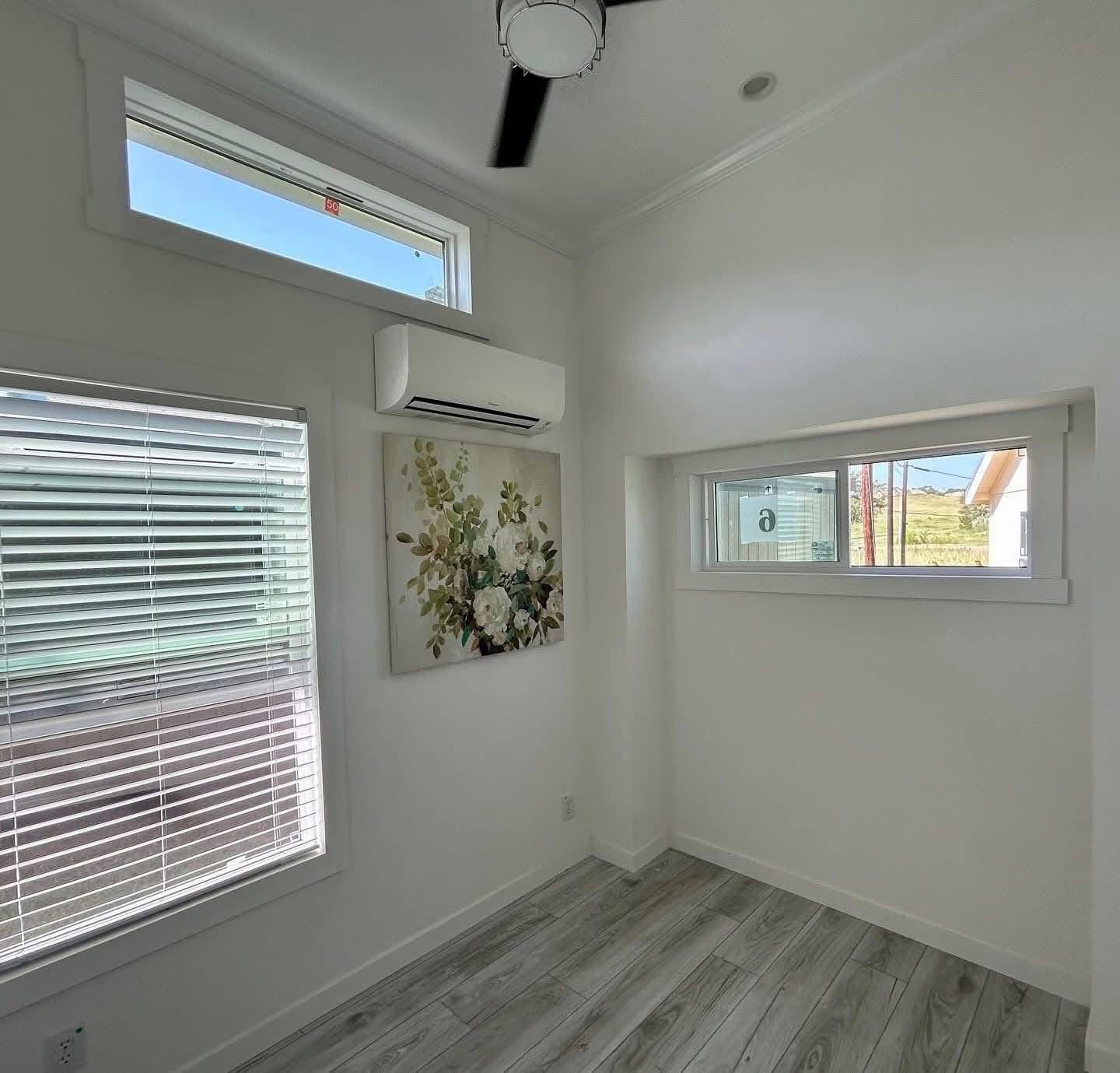 Minimalist room with light gray wooden floors, white walls, and a wall-mounted air conditioner. It features floral artwork and horizontal windows.