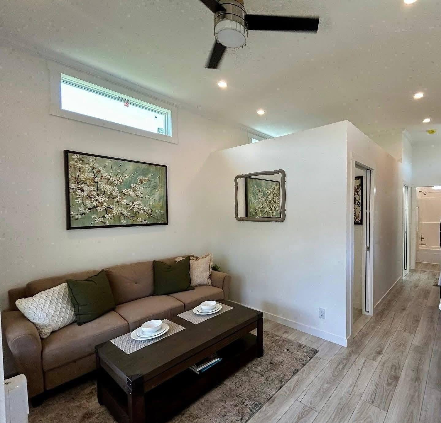 A cozy living room with a taupe sofa adorned with green and white cushions, a dark wood coffee table set with plates, and floral wall art. Light wood flooring and a ceiling fan add to the modern, inviting atmosphere.