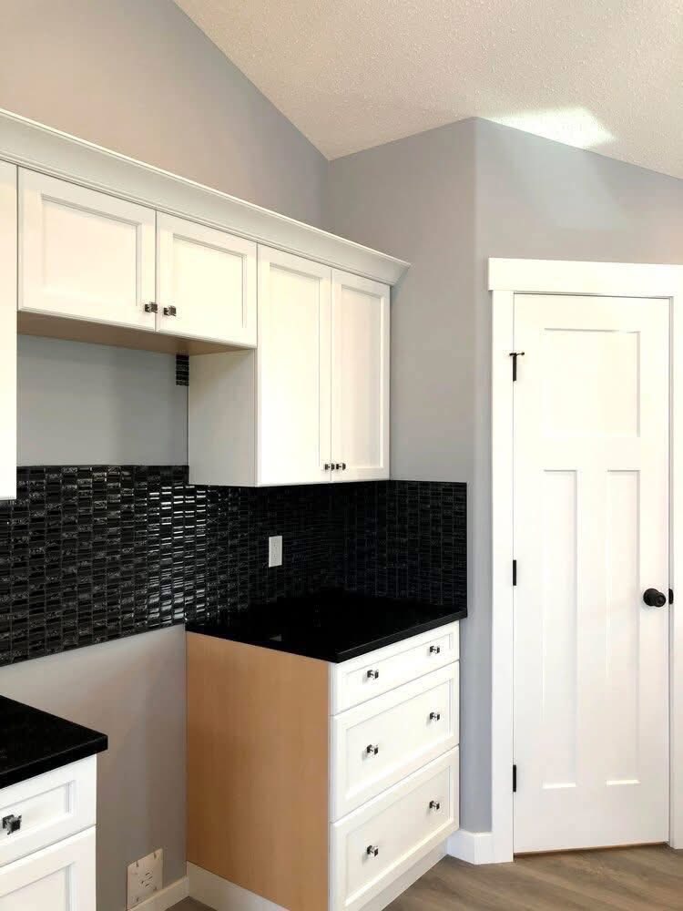 Modern kitchen with white cabinets, black countertops, and black tile backsplash. Light gray walls and a white door add a clean, minimalistic feel.
