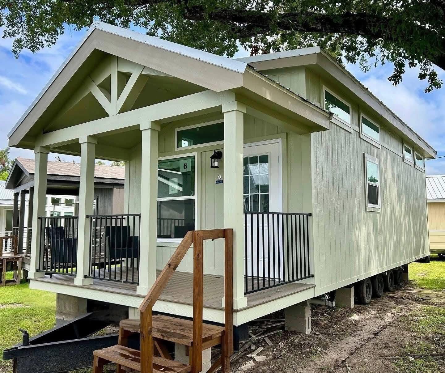 A small green mobile home with a front porch featuring wooden steps and black railings. Large windows and a gabled roof give a cozy, inviting look.