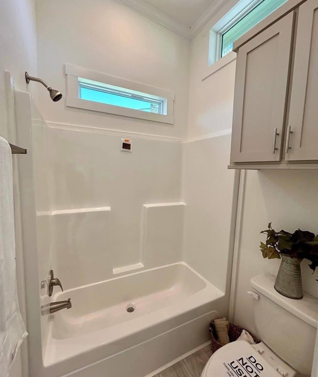 Bright bathroom with a white bathtub, overhead shower, and small window. Gray cabinet above the toilet with decorative plant; modern and clean.