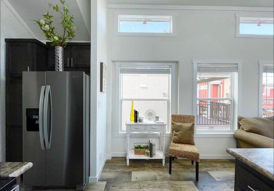 Modern room with a stainless steel fridge, white side table, and wicker chair by large windows. Bright, airy ambiance with simple decor.