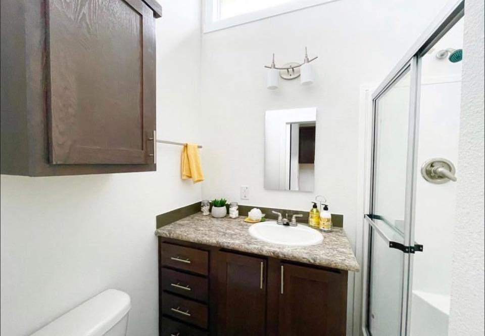 Compact bathroom with dark wood cabinets, a marble countertop, and a round sink. A yellow towel hangs on a rack. A glass shower door and overhead lighting add a modern touch.
