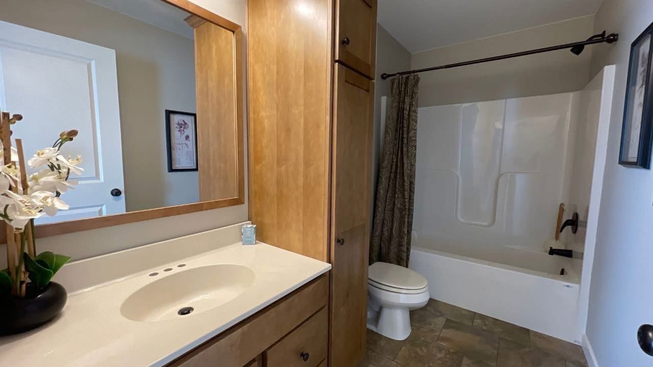 A warm bathroom with wooden cabinets, white sink, large mirror, and a vase with white orchids. Bathtub with patterned curtain and toilet nearby.