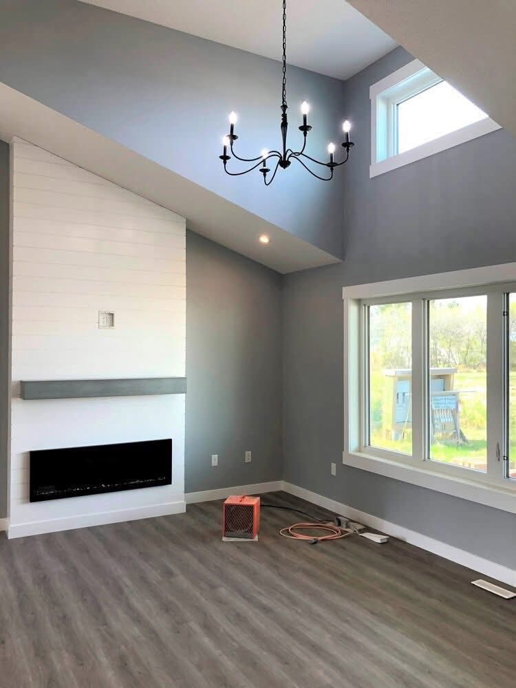 Modern living room with a high ceiling, featuring a black chandelier, gray walls, large window, electric fireplace, and light wood flooring. Cozy and bright.