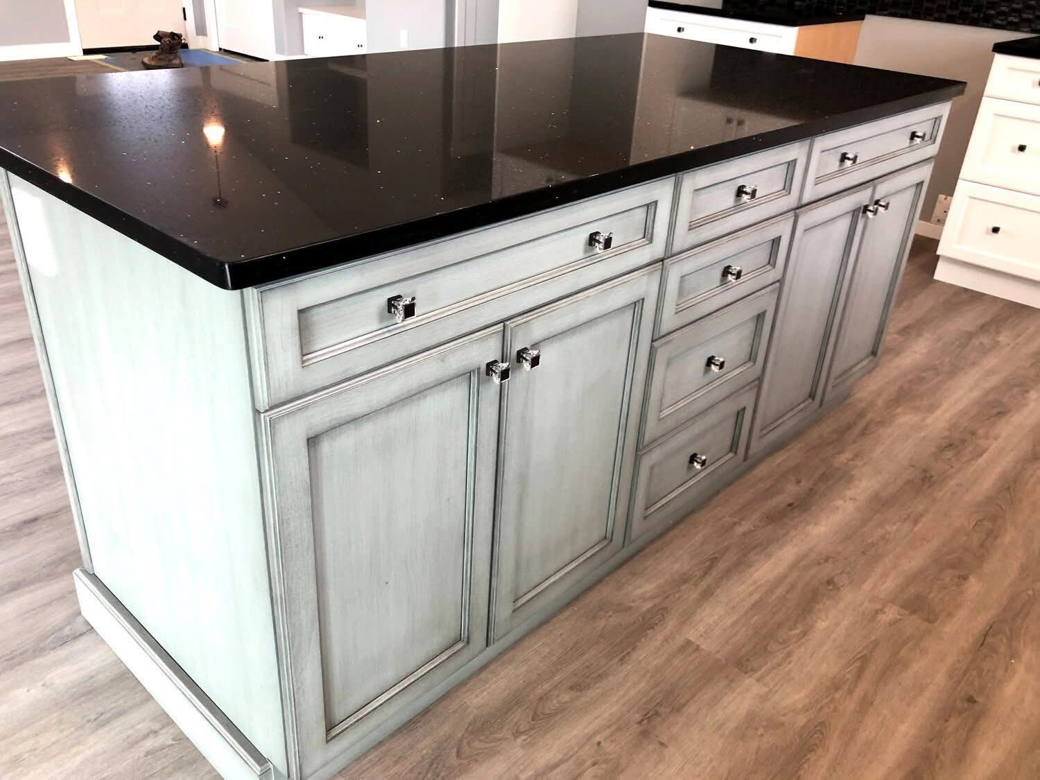 A modern kitchen island with a glossy black countertop and light grey cabinetry. It features multiple drawers and cabinets, set on wood flooring.