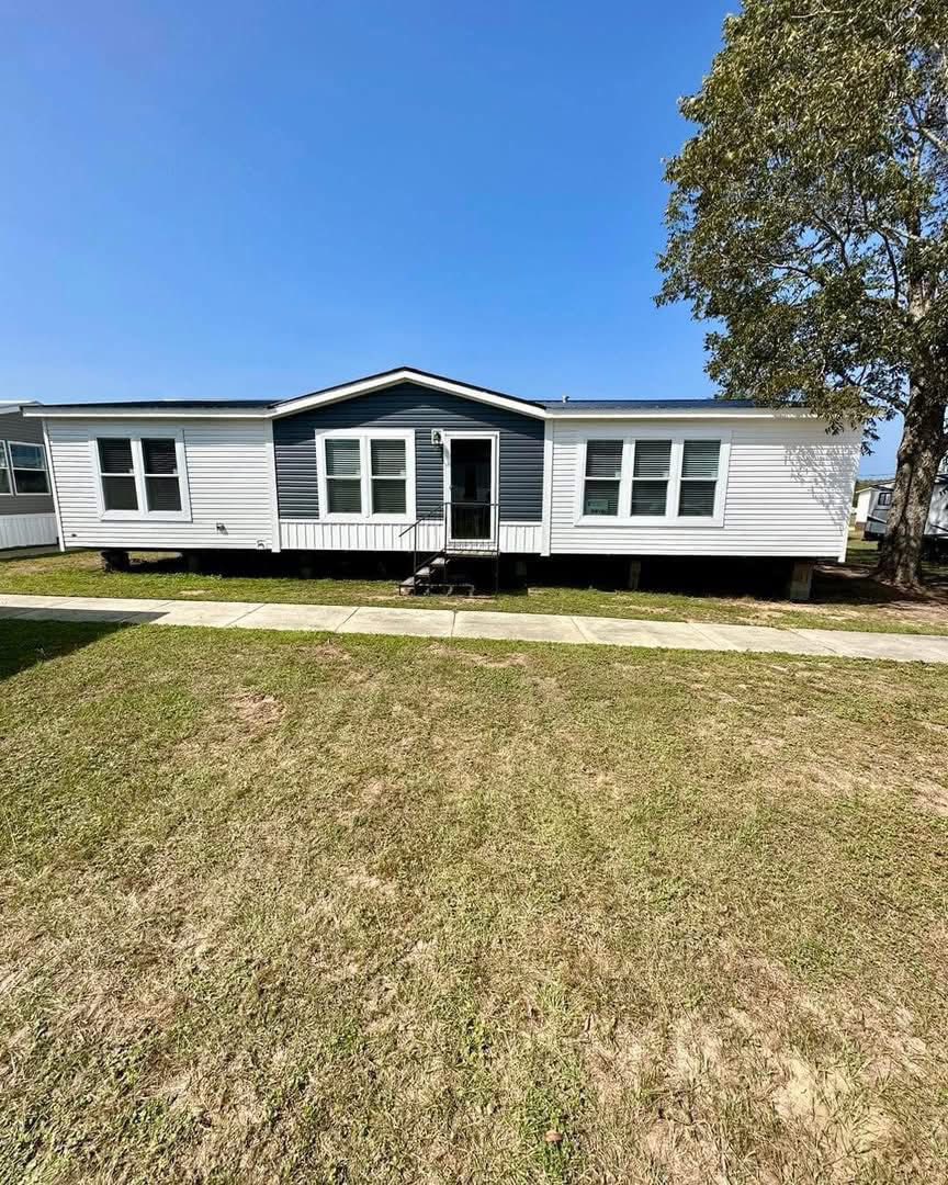 Single-story white mobile home with a dark blue accent, set on a clear day. A concrete path leads to the front steps, with grass in the foreground. A large tree stands to the right.