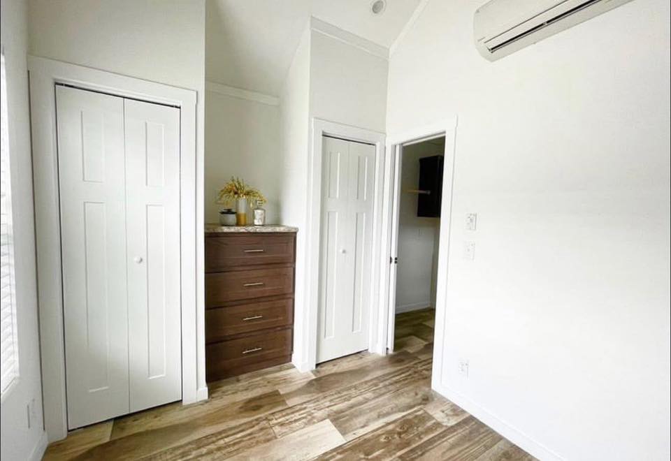 A small, bright room with wooden floors and white walls. It features two closed white doors, a dark wooden chest of drawers with decor on top, and a wall-mounted air conditioner, conveying a neat and minimalist atmosphere.