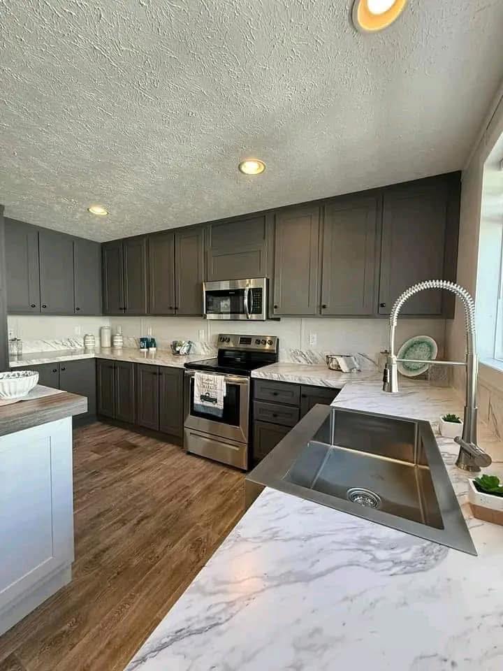 Modern kitchen with dark cabinets, marble countertops, and stainless steel appliances. Large sink with a tall faucet, wooden floor, and bright overhead lighting.